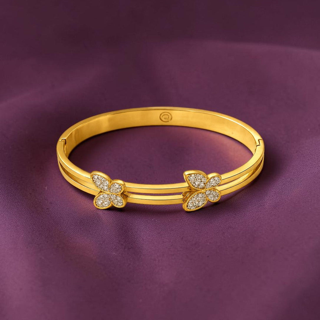 A polished yellow gold hinged bangle bracelet with two butterfly motifs, one adorned with small clear pavé stones, displayed on a draped purple fabric background.