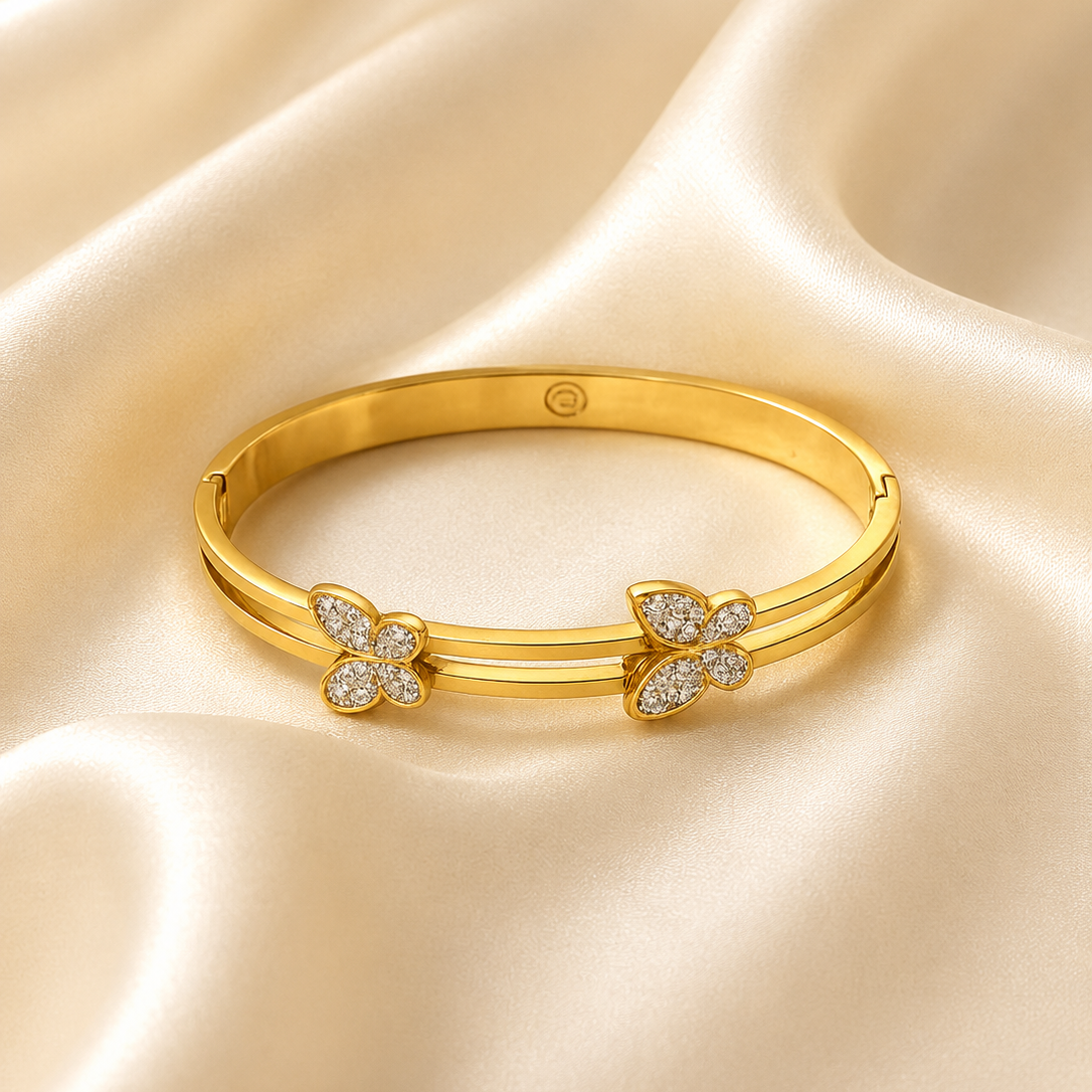 A gold bangle bracelet with two butterfly motifs paved with small clear stones, displayed on a draped cream satin fabric background.