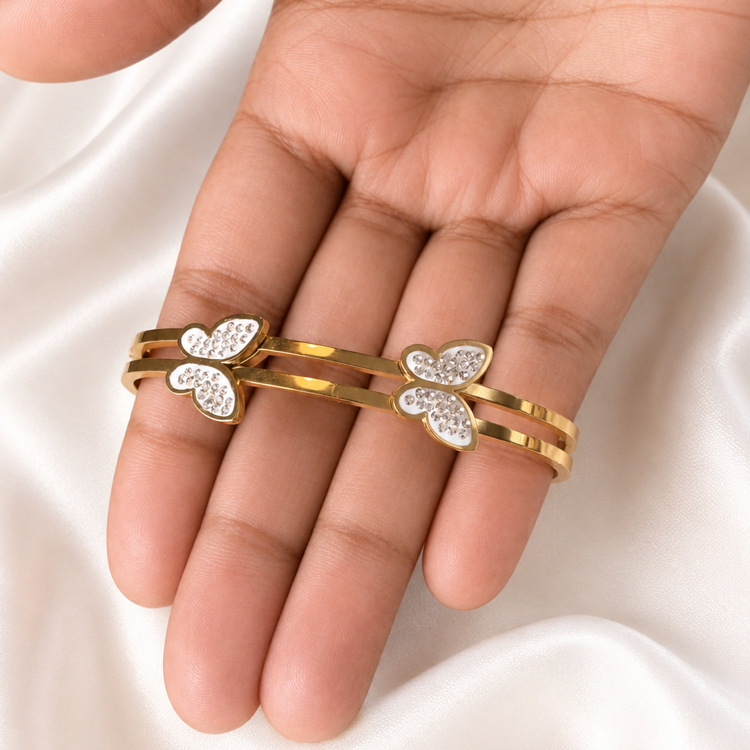 A close-up of a hand wearing three gold rings, each decorated with a small heart-shaped cluster of clear rhinestones, set against a soft white satin fabric background.