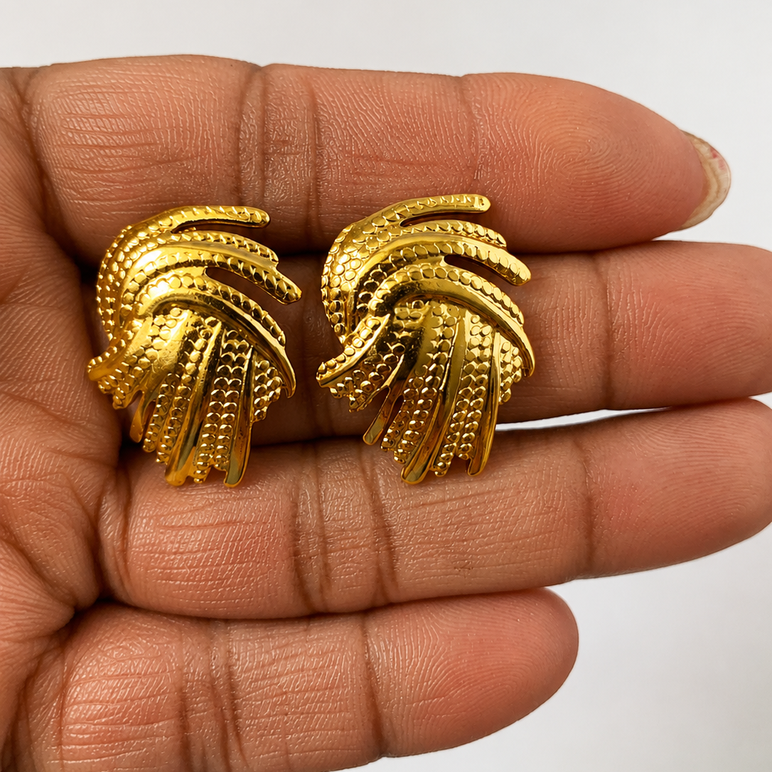 Gold tone earrings held between fingers against a neutral background