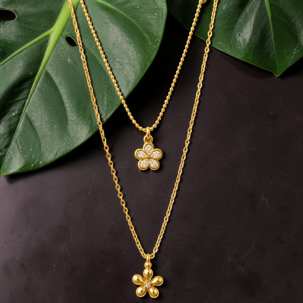 Gold tone necklaces with flower pendants on a dark surface with green leaves.