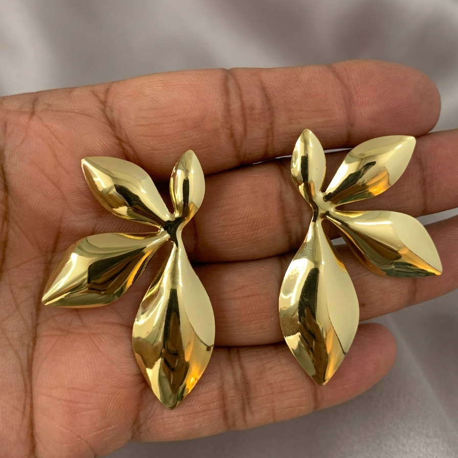Gold leaf-shaped earrings held in a hand against a neutral background