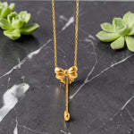 Gold tone bow-shaped necklace on a black marble surface with green succulents.
