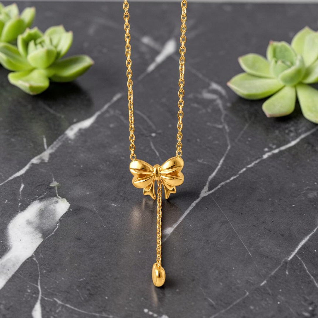 Gold tone bow-shaped necklace on a black marble surface with green succulents.