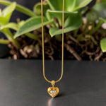 Gold tone necklace with a heart-shaped pendant on a dark surface with green leaves in the background