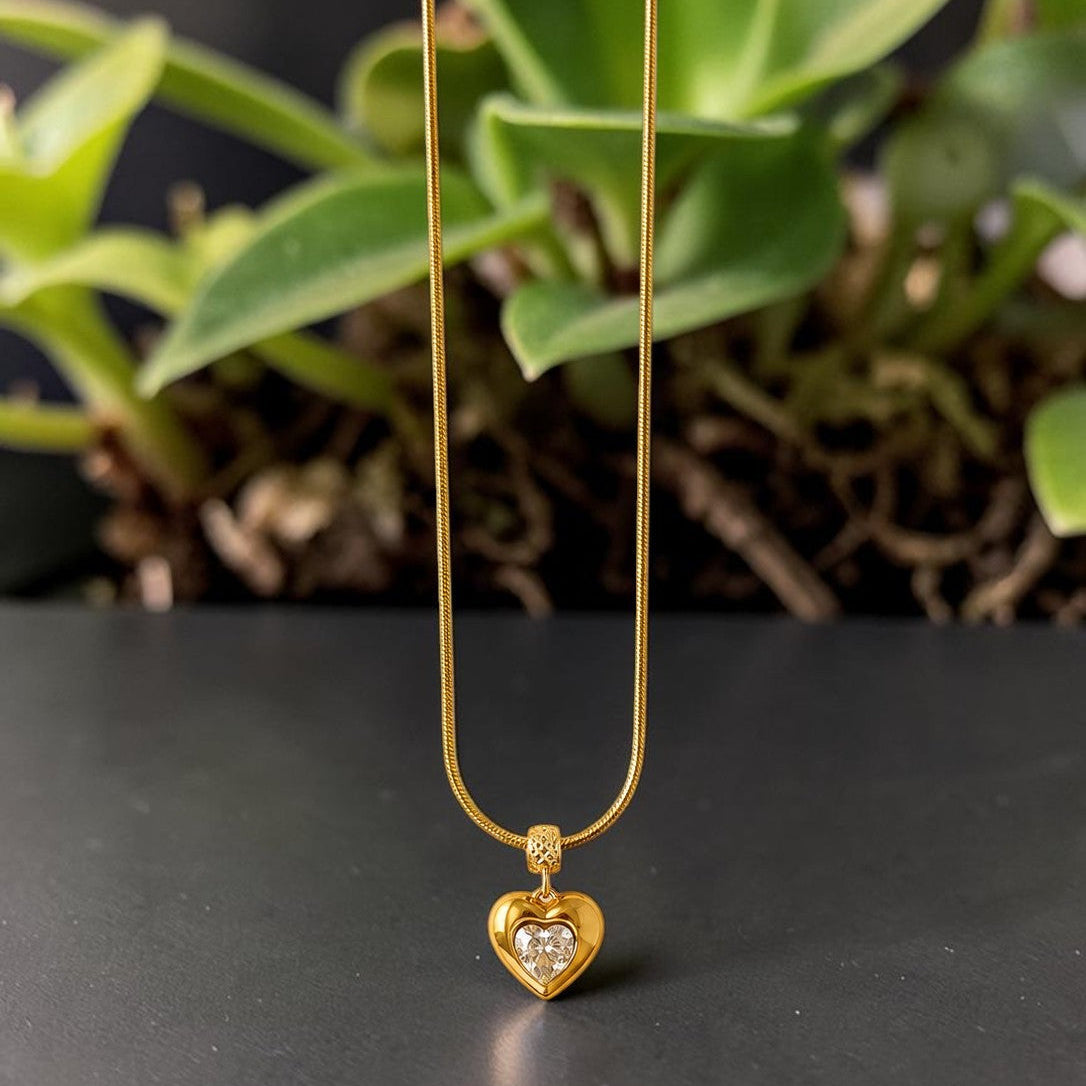 Gold tone necklace with a heart-shaped pendant on a dark surface with green leaves in the background