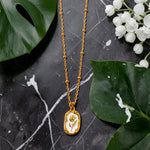 Gold tone necklace with a pendant on a dark surface with green leaves and white flowers.