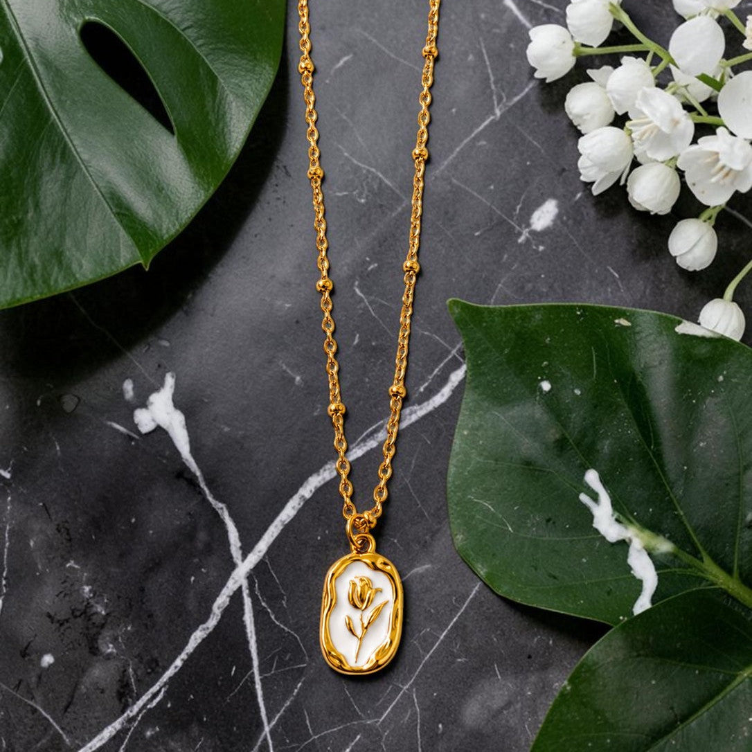 Gold tone necklace with a pendant on a dark surface with green leaves and white flowers.