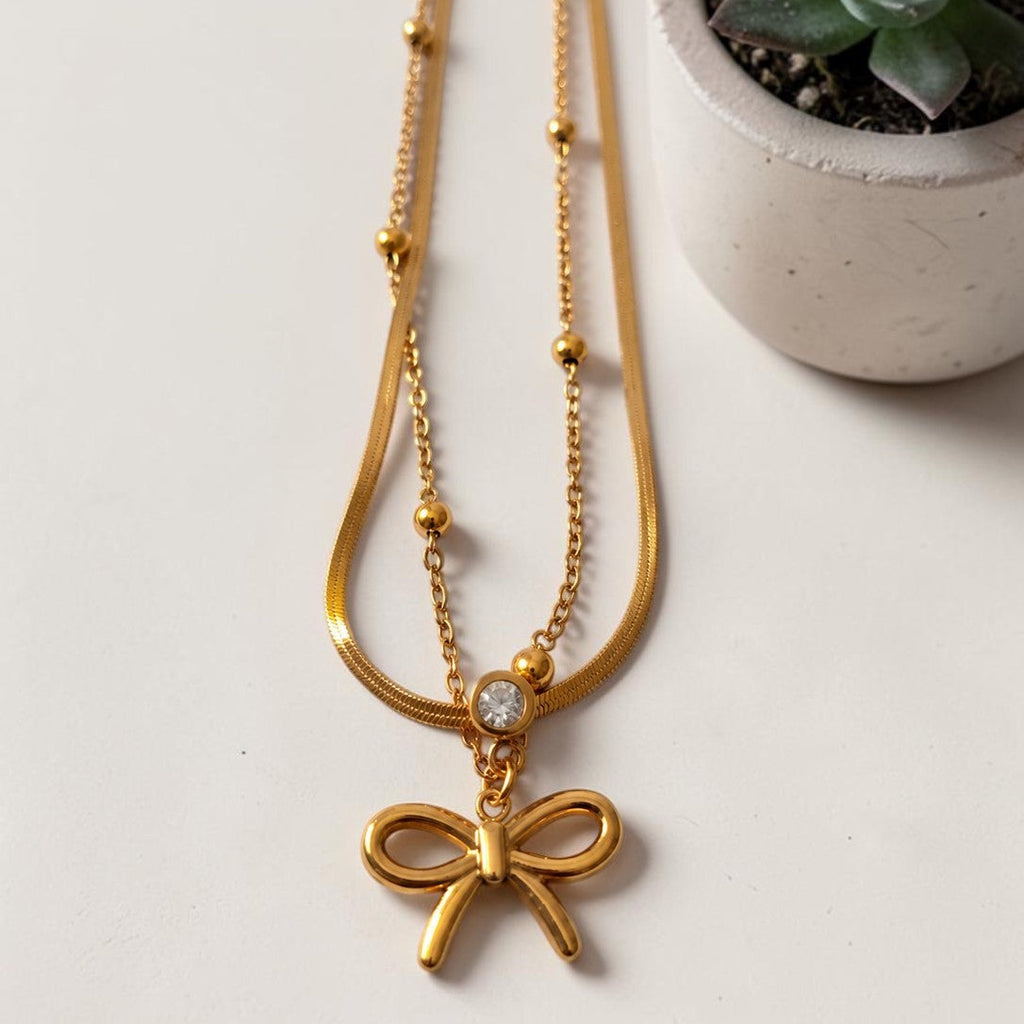 Gold tone necklace with a bow pendant on a light surface with a small plant in the background