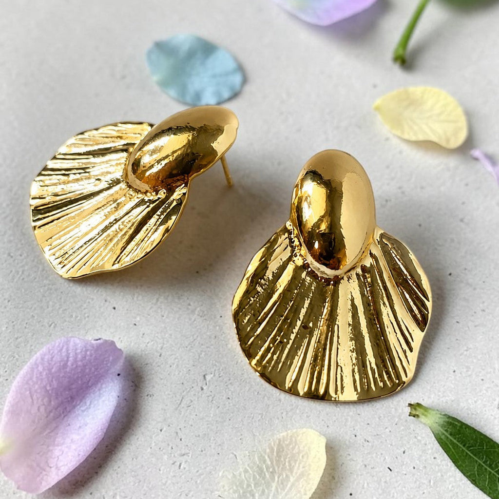 Gold shell-shaped earrings on a light surface with flower petals and leaves.