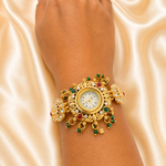 An ornate gold-toned wristwatch with red and green gemstone accents, shown worn on a person's wrist against a draped satin fabric background.