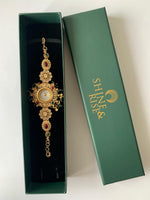 An ornate gold-toned traditional Indian bracelet with red and green stone accents, displayed inside an open dark green box next to its closed lid, which features the brand name 'SHREE RISES' in gold.