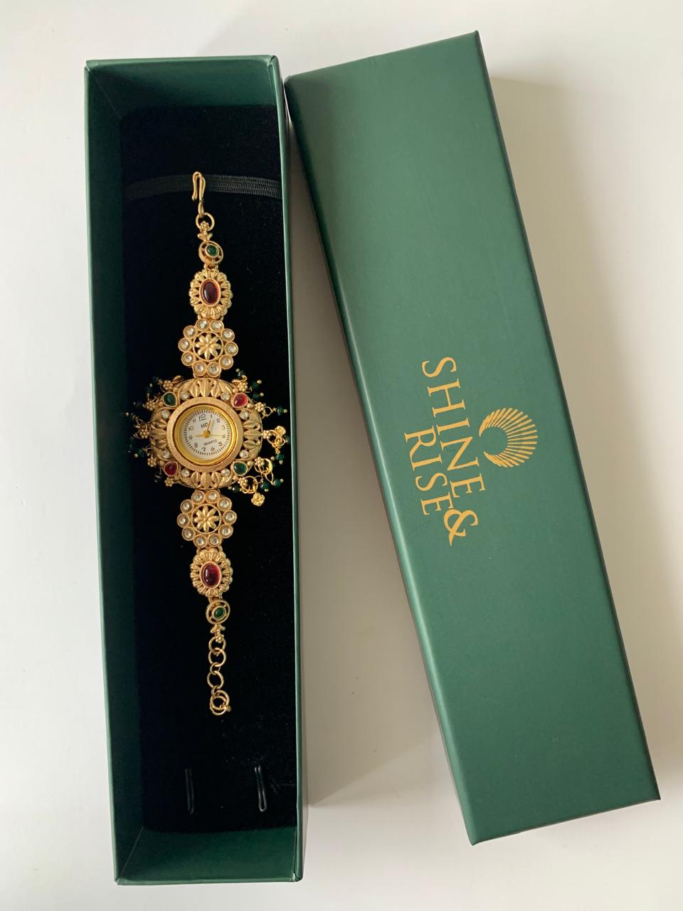 An ornate gold-toned traditional Indian bracelet with red and green stone accents, displayed inside an open dark green box next to its closed lid, which features the brand name 'SHREE RISES' in gold.