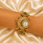 An ornate gold-toned bangle watch with green and red enamel details, worn on a wrist against a draped champagne satin background.