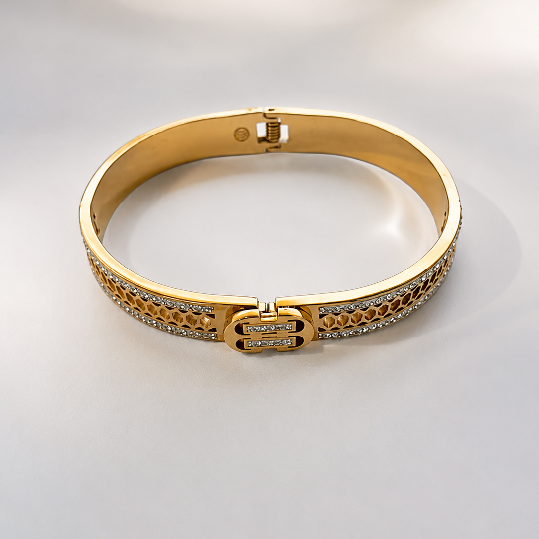 A gold bangle bracelet with pavé-set clear stones and a central interlocking double-helix clasp, photographed on a light gray background.