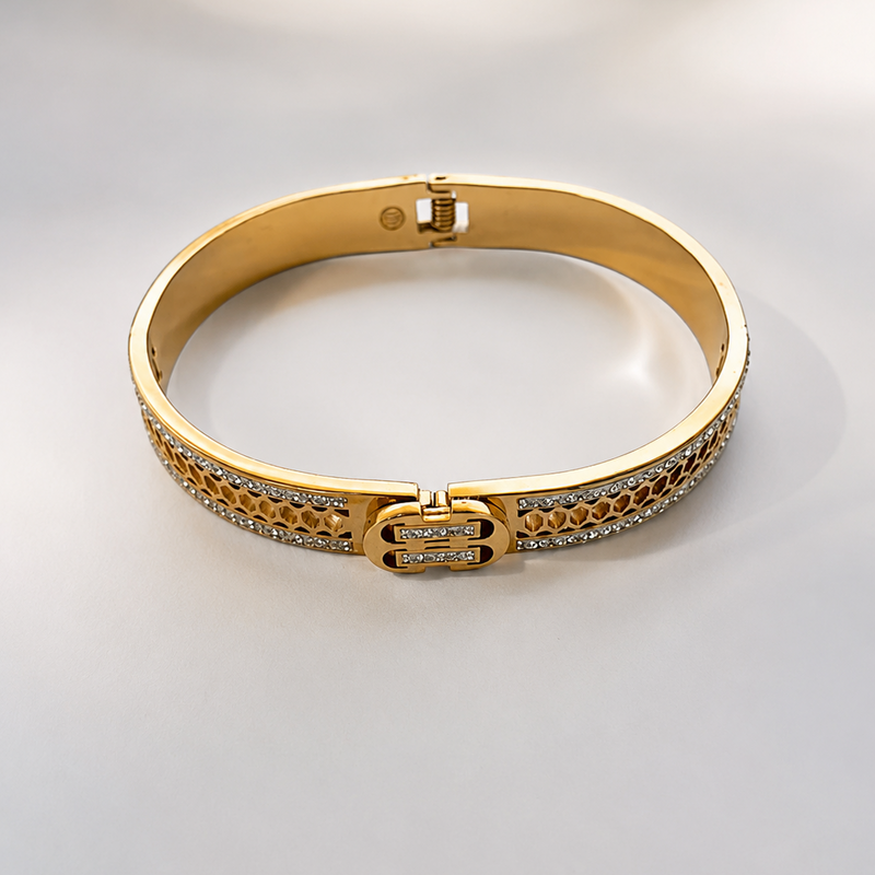 A gold bangle bracelet with pavé-set clear stones and a central interlocking double-helix clasp, photographed on a light gray background.