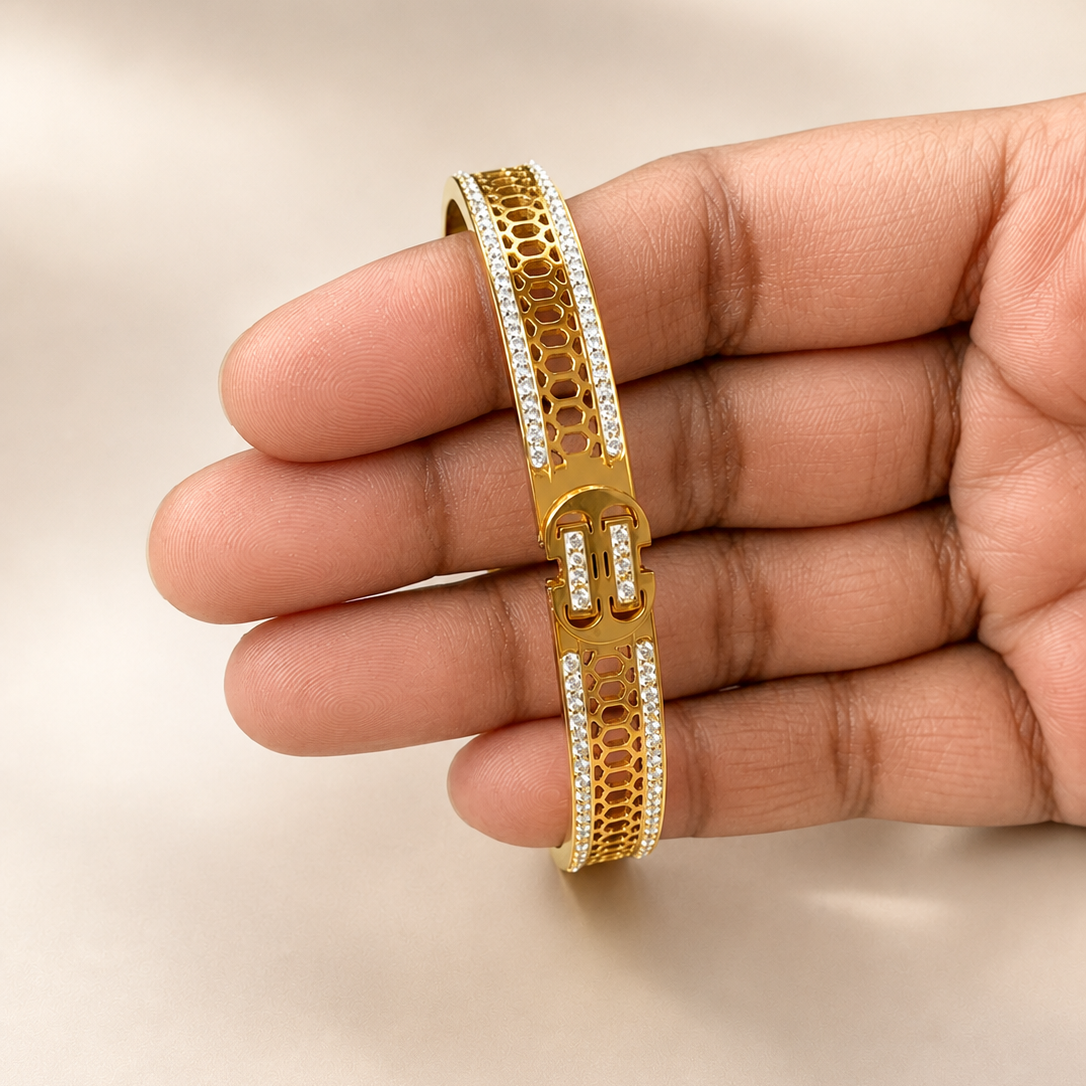 A gold-toned bangle bracelet with textured honeycomb patterns and rows of small clear stones, held on a person's fingers against a soft beige background.