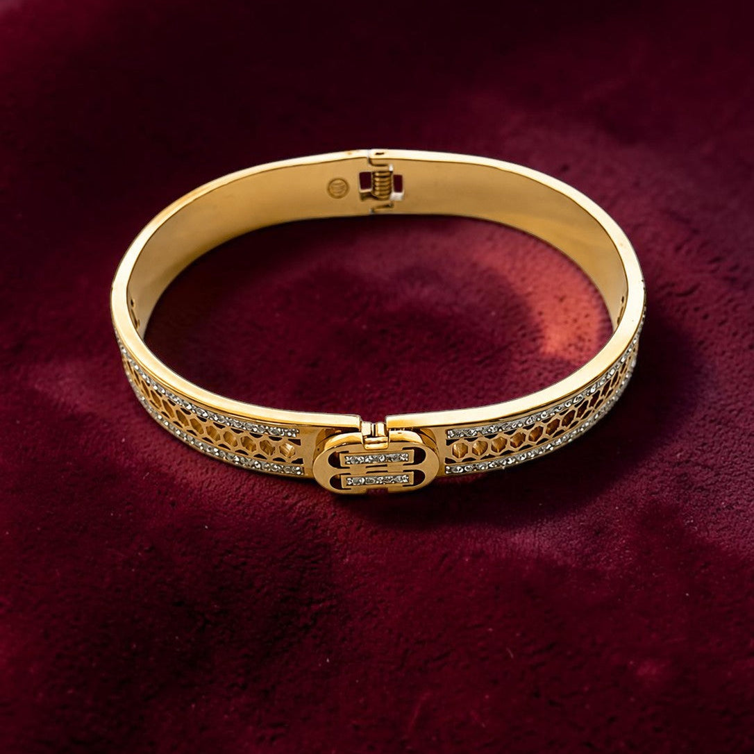 A gold bangle bracelet with pavé-set clear stones and a central clasp, displayed on a deep burgundy velvet background.