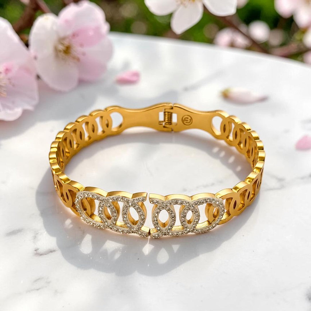 A gold bangle bracelet with a repeating interlocking chain-link design, featuring a front section paved with small clear stones. The bracelet is displayed on a white marble surface with soft pink blossoms blurred in the background.