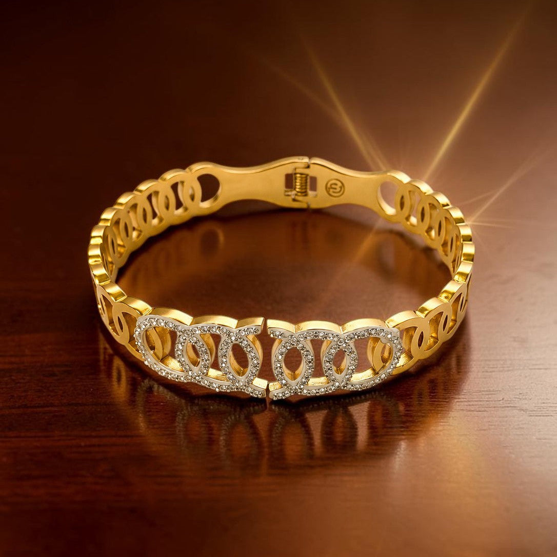 A gold-toned bracelet with repeating interlocking links and a front section set with clear stones, displayed on a dark brown reflective surface.