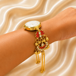 A gold-toned wristwatch with a decorative charm featuring red and gold details, shown worn on a person's wrist against a draped satin fabric background.