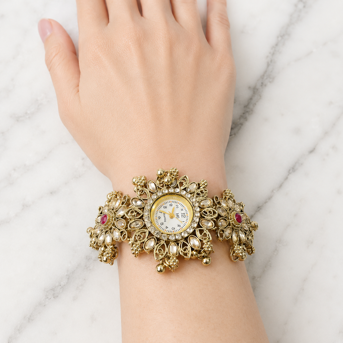 An ornate gold-toned bracelet watch with pearl-like beads, small red gemstones, and a round white dial, worn on a person's wrist against a light marble background.