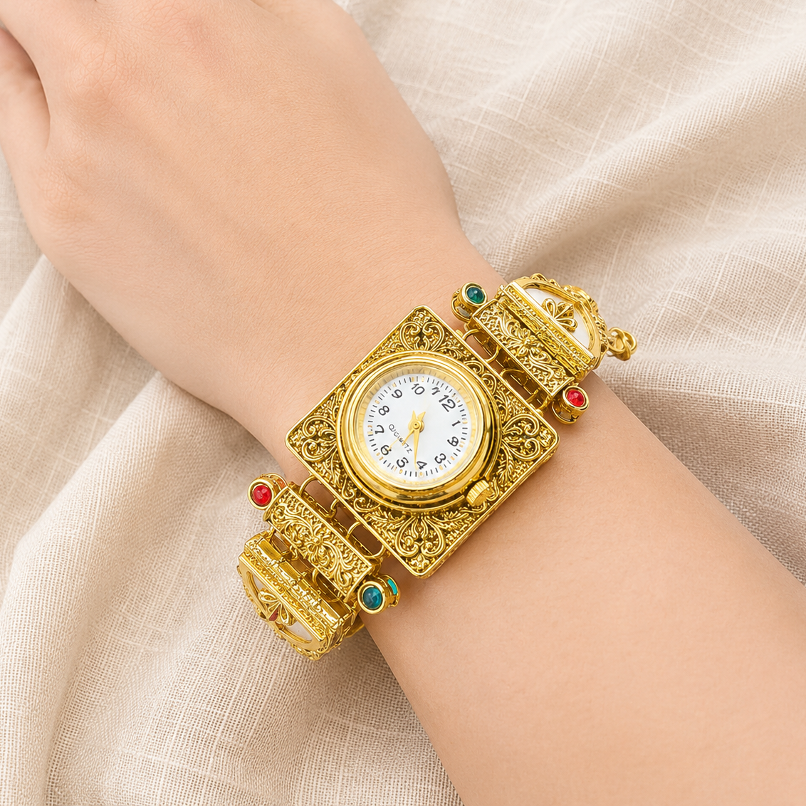 A close-up of a person's wrist wearing an ornate gold-toned watch with a square dial, decorative filigree bands, and small red and green gemstone accents, set against soft beige fabric.