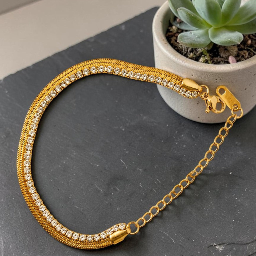 Gold tone necklace with diamond accents on a dark surface next to a potted plant
