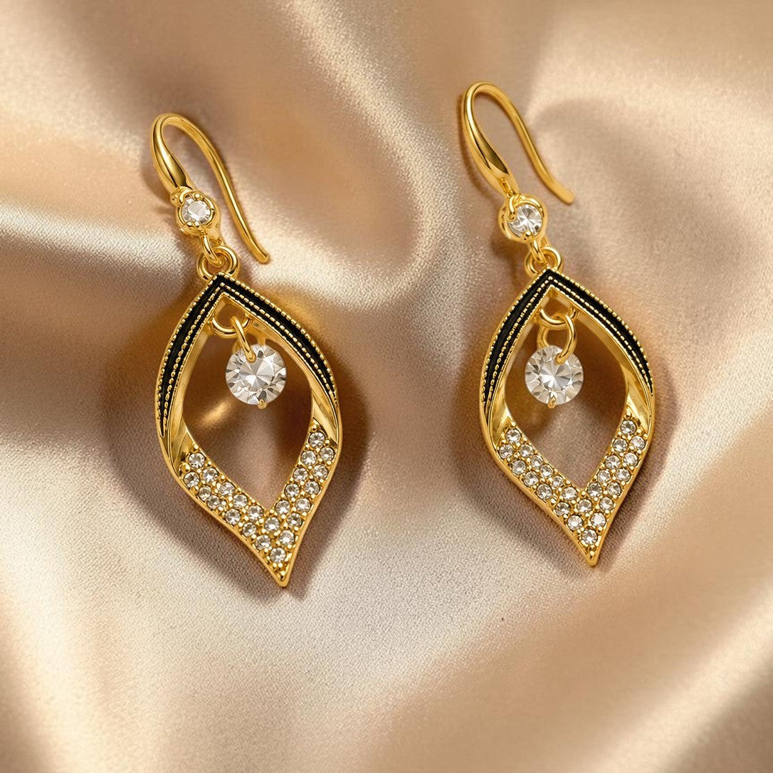 A pair of gold drop earrings with a teardrop-shaped openwork design, accented with black enamel, pavé-set clear crystals, and a central round clear stone, displayed on a beige satin fabric background.