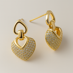Gold tone heart-shaped earrings with diamond-like stones on a beige background