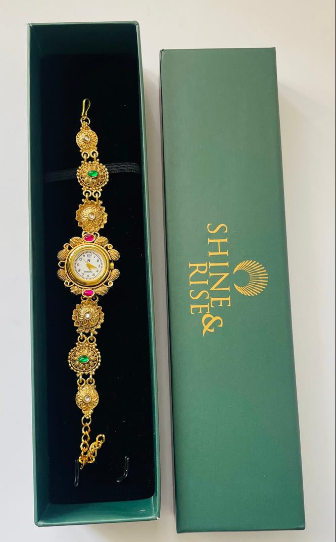 A gold-plated traditional Indian watch bracelet with green and red stone accents and a central watch face, displayed inside an open dark green presentation box next to its closed box which features the brand name 'SHINE RISE'.