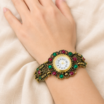 A close-up of a person's hand and wrist wearing an ornate vintage-style watch with a gold-toned bracelet, green and red gemstone accents, and a white dial.