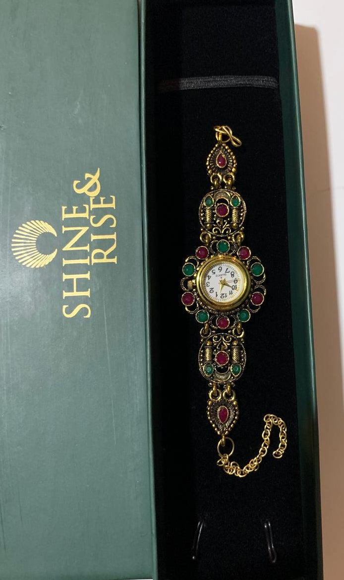A gold-tone vintage-style watch bracelet with red and green enamel details, displayed inside a dark green Shine & Rise gift box.