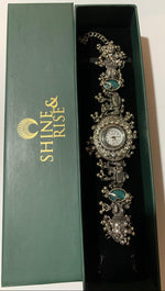 An ornate silver-toned watch with turquoise green stones, presented inside a black gift box next to its green Shine & Rise brand packaging.