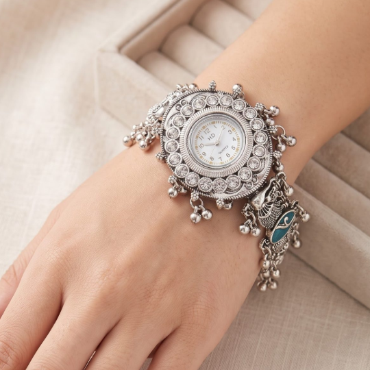 A close-up of a woman's hand and wrist wearing an ornate silver-toned watch with crystal embellishments and a dangling owl charm, set against a soft beige textured background.