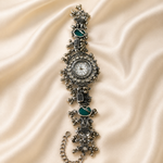 An ornate silver-tone watch with a chain strap, featuring green enamel details and clear rhinestones, displayed on a draped cream satin fabric background.
