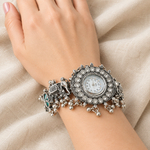A close-up of a woman's wrist wearing an ornate silver-toned watch with a circular dial, rhinestone-embellished bezel, elephant motif, and dangling bead chains, set against draped beige fabric.