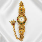 An ornate gold-plated traditional Indian wrist watch with a chain strap, decorated with red and green enamel, a white dial, and small stones, displayed on a white satin background.