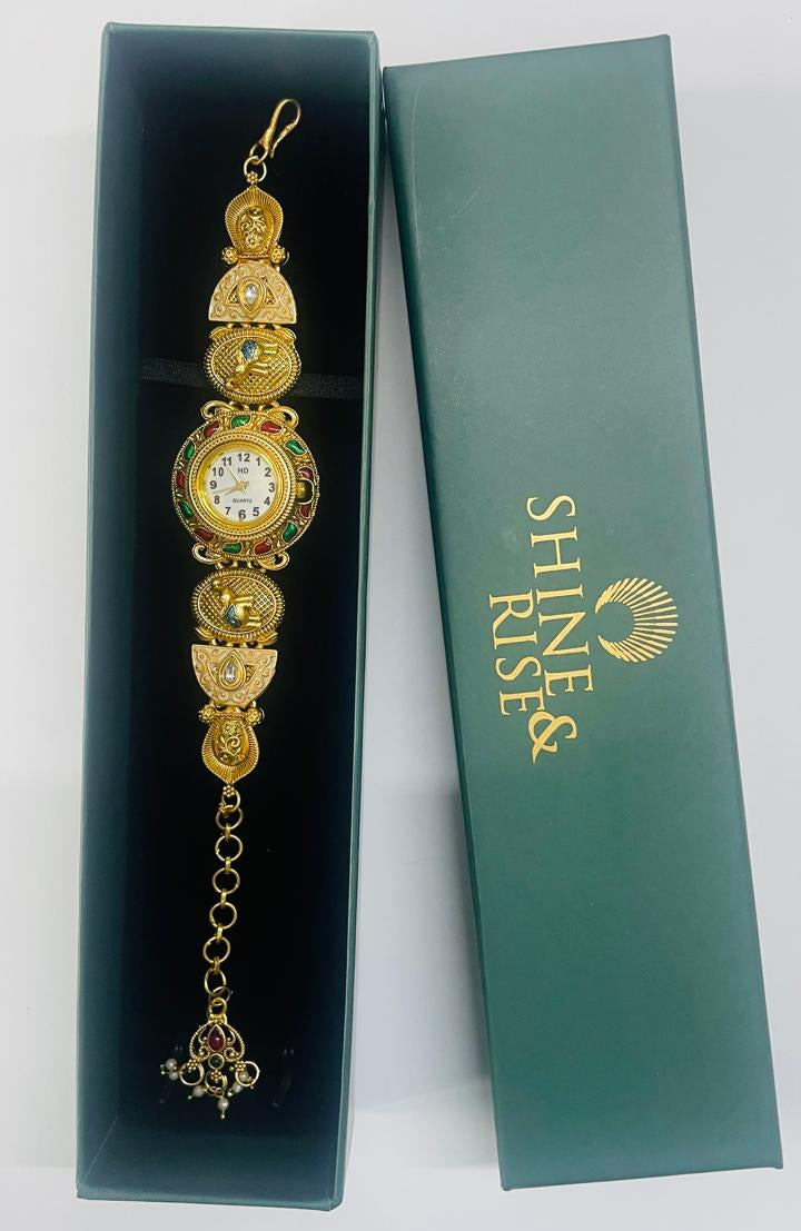 A gold-toned ornate watch with a white dial and small colored gem accents, displayed inside an open dark teal presentation box next to a closed matching box printed with the brand name 'SHINE RISE'.