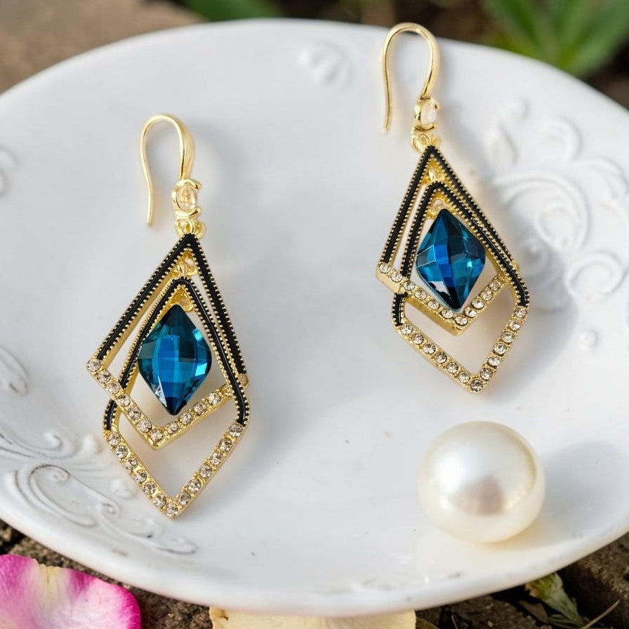 Golden earrings with blue gemstones on a white dish with a pearl and pink petals.
