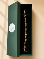 A gold-toned bracelet with alternating pink and purple gemstone accents displayed inside an open dark green presentation box with a white circular logo.