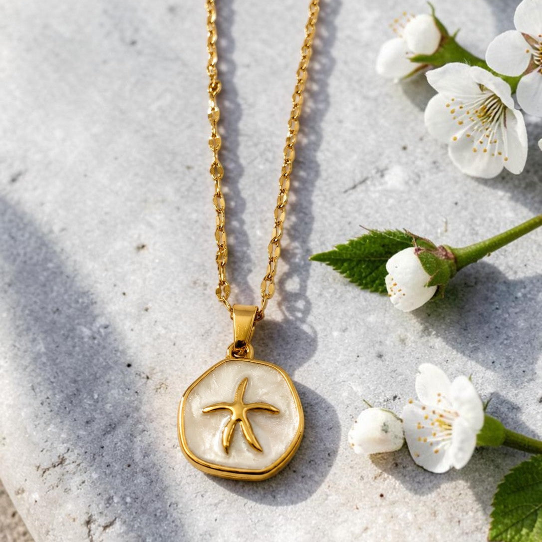 Gold tone necklace with a starfish pendant on a stone surface with white flowers and green leaves.