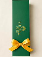 A tall, dark green rectangular gift box with gold 'SHINE & RISE' branding and a gold shell logo, tied with a bright yellow satin ribbon bow at the bottom, on a light beige background.