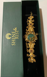 An ornate gold-toned traditional jewelry piece with a green oval centerpiece and small red stone accents, displayed inside an open dark green Shine & Rise gift box.