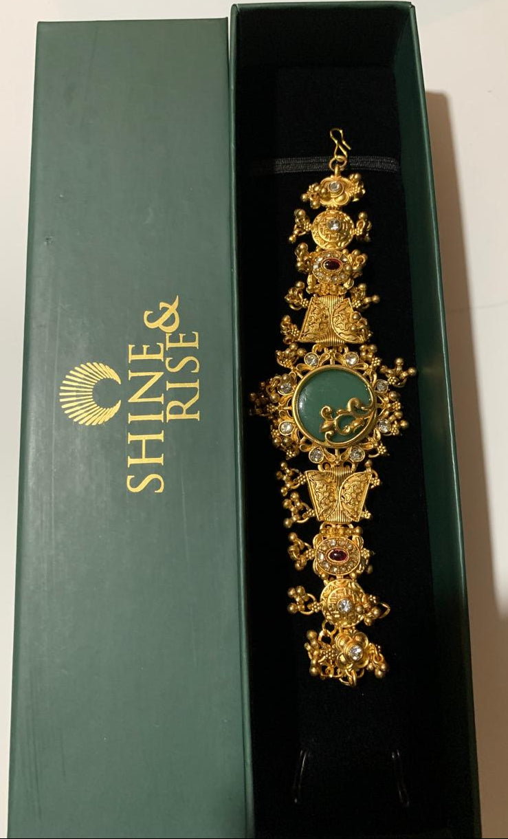 An ornate gold-toned traditional jewelry piece with a green oval centerpiece and small red stone accents, displayed inside an open dark green Shine & Rise gift box.
