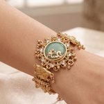 A close-up of a person's wrist wearing an ornate gold-toned bracelet with a round light green stone centerpiece and decorative filigree details.