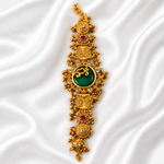 An ornate gold-toned traditional bracelet or amulet featuring a large green oval stone at the center, surrounded by red and clear gemstone accents, displayed on draped white satin fabric.