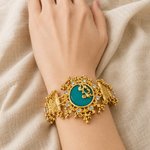 A close-up of a person's wrist wearing an ornate gold-toned bracelet with a round turquoise-blue center stone and small clear stones, set against draped beige fabric.