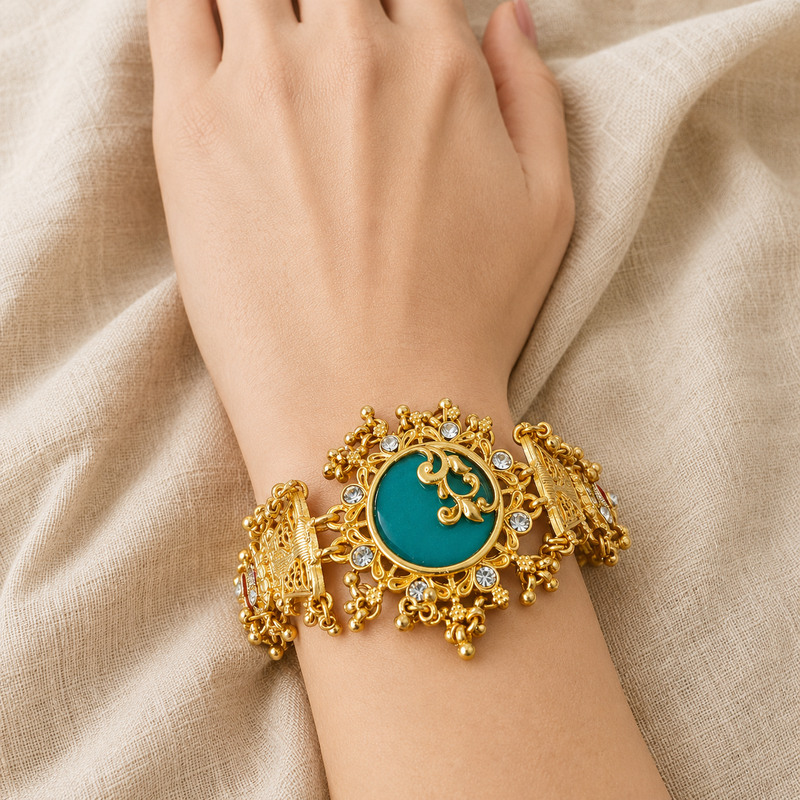 A close-up of a person's wrist wearing an ornate gold-toned bracelet with a round turquoise-blue center stone and small clear stones, set against draped beige fabric.