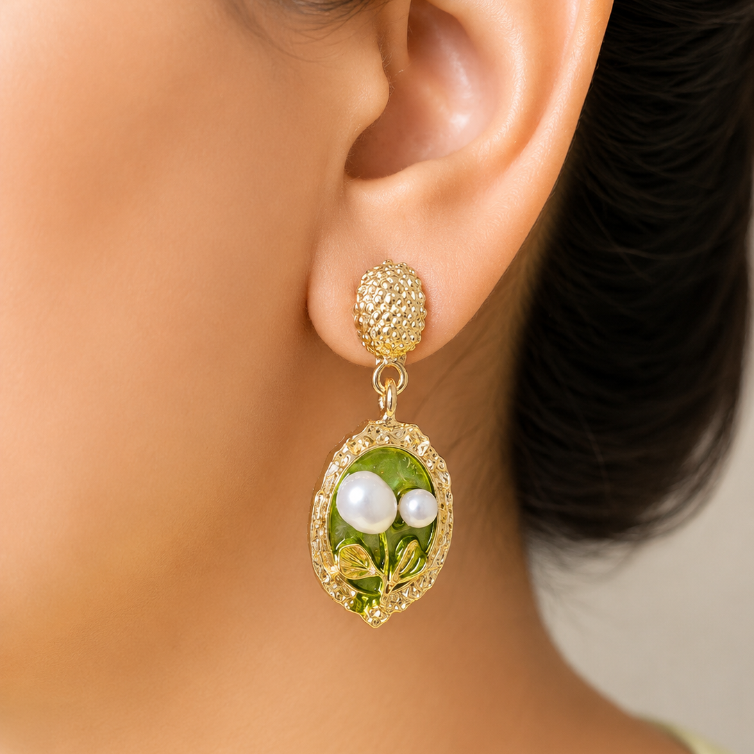 A close-up of a model's ear wearing a gold drop earring with a textured top and an oval green leaf and pearl charm.
